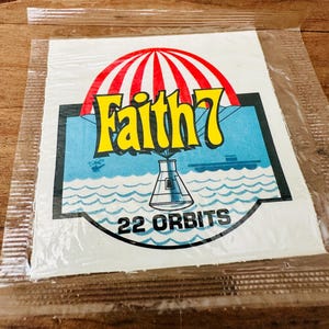 May include: A vintage sticker in a clear plastic sleeve. The sticker features the text "Faith 7" in yellow, a red and white striped parachute, and a blue background with a rocket and the text "22 ORBITS".