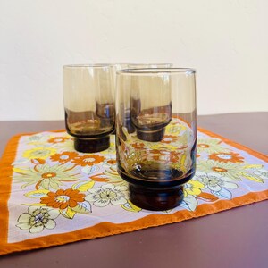 May include: Four vintage brown-tinted drinking glasses with a dark brown base. The glasses are cylindrical and sit on a floral patterned cloth with orange, yellow, and white flowers. The cloth has an orange border.