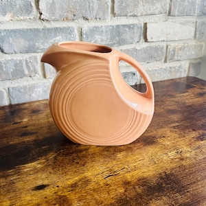 May include: A peach-colored ceramic pitcher with a circular design and a large handle. The pitcher has a spout for pouring and sits on a wooden surface. The background features a brick wall.