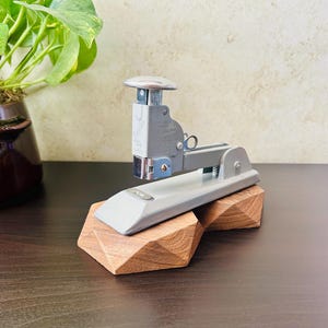 May include: A gray metal stapler rests on a geometric wooden base. The stapler has a silver top and a long, flat base. The wooden base is a warm brown color and has a faceted design. A green plant is in the background.