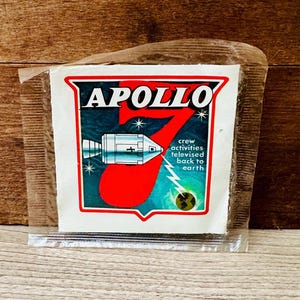 May include: A vintage Apollo 7 sticker in a clear plastic sleeve. The sticker features a red number 7, a spacecraft illustration, and the text "APOLLO" and "crew activities televised back to earth" against a blue and green background.