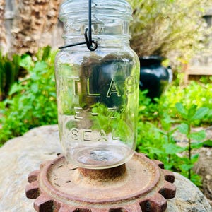 May include: A clear glass jar with a metal lid and a wire handle. The jar has the words "ATLAS E-Z SEAL" embossed on the side. The jar is sitting on a rusty metal gear.