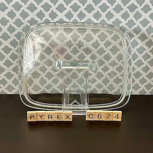 May include: A clear glass baking dish with a lid and a handle. The dish is sitting on a brown surface with the words "PYREX" and "C624" spelled out in letter tiles.