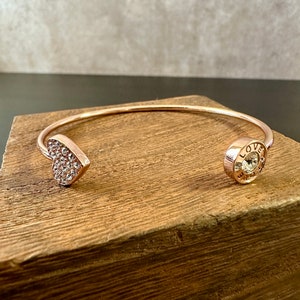 May include: Rose gold-toned cuff bracelet. One end features a heart shape encrusted with small, clear crystals. The other end has a circular charm with the word "LOVE" and a central crystal. The bracelet is displayed on a wooden surface.