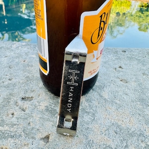 May include: A silver bottle opener with the word "HANDY" printed vertically. The opener is next to a brown glass bottle with a yellow and white label. The bottle is partially visible, with the opener in the foreground.