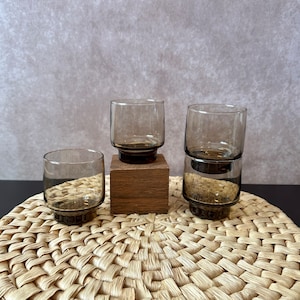 May include: Four clear, short, gray-tinted drinking glasses. The glasses have a dark brown base and are stacked on a wooden block and a woven mat. The glasses are empty and appear to be made of glass.