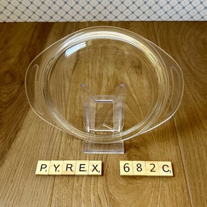 May include: A clear Pyrex glass lid with two side handles, resting on a clear plastic stand. The lid is circular with a slightly raised edge. The word "PYREX" and the number "682 C3" are spelled out with wooden tiles.
