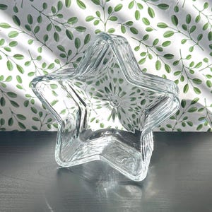 May include: A clear glass star-shaped dish with a slightly rippled texture. The dish is empty and sitting on a dark gray surface.