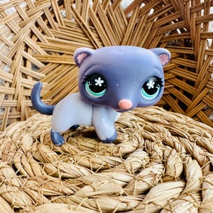 May include: A collectible toy figurine of a cat with a lavender head and tail, and a white body. The cat has large, expressive eyes with a flower design, a pink nose, and pink inner ears. The figurine is set against a woven background.