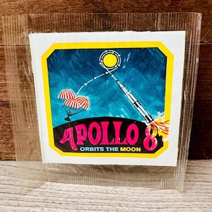 May include: A vintage Apollo 8 sticker, featuring a rocket launching towards the moon, with three parachutes and the text "Apollo 8 Orbits the Moon." The sticker is encased in a clear plastic sleeve.