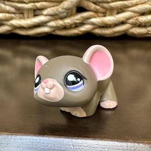 May include: A small, brown toy mouse with large, black eyes and pink ears. The mouse has a light pink nose and a white mouth. The toy is made of a hard plastic material and is sitting on a dark brown surface.