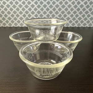 May include: Four clear glass bowls stacked on a dark surface. Each bowl has a decorative embossed design around the rim and a central motif. The bowls are transparent, allowing the background to be partially visible.