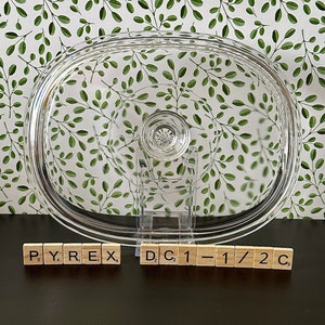 May include: A clear glass Pyrex baking dish lid with a knob on top. The lid is sitting on a dark surface with the words "PYREX" and "DC 1-1/2 C" spelled out in letter tiles.