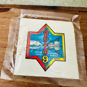 May include: A vintage Apollo 9 sticker in a clear plastic sleeve. The sticker features a star-shaped design with the words "APOLLO 9" and illustrations of the lunar module and spacecraft. The sticker is in a clear plastic sleeve.