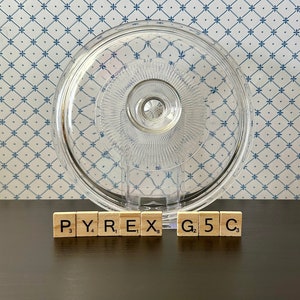May include: A clear glass Pyrex lid with a textured surface. The lid is sitting on a dark brown surface with the word "PYREX G5C" spelled out in letter tiles.