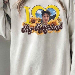 May include: Cream-colored sweatshirt with a graphic design. The design features the words "I ❤️ My Boyfriend" in yellow and brown, with a photo of a man inside a yellow heart shape, surrounded by sunflowers.