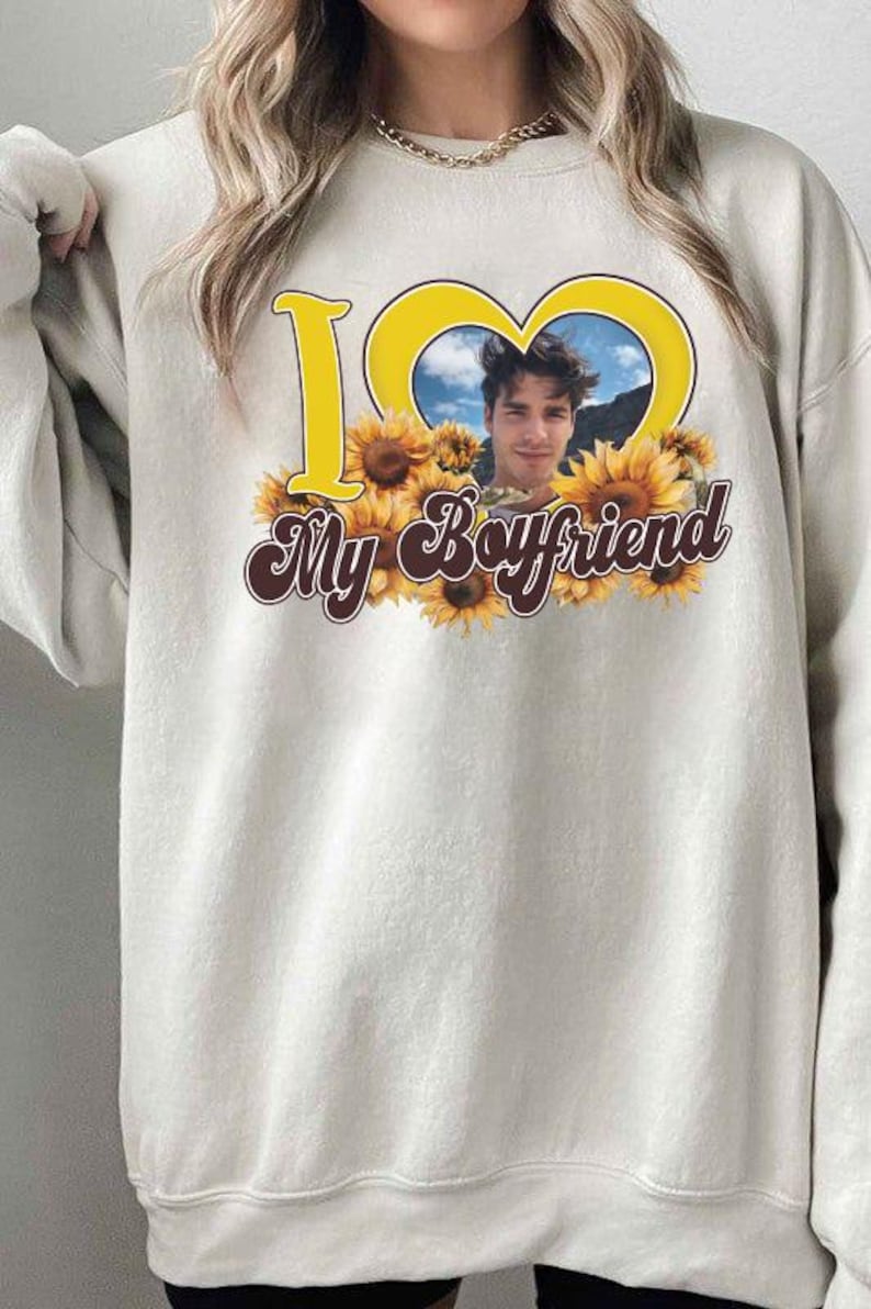 May include: Cream-colored sweatshirt with a graphic design. The design features the words "I ❤️ My Boyfriend" in yellow and brown, with a photo of a man inside a yellow heart shape, surrounded by sunflowers.