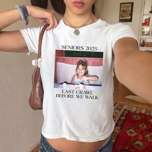 Op de afbeelding: Wit crop top met de tekst "Seniors 2025" en "Last Crawl Before We Walk" erop gedrukt. Een foto van een jong meisje is gecentreerd op het shirt.