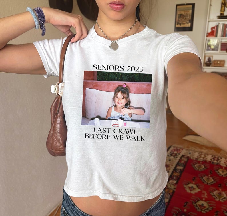 Op de afbeelding: Wit crop top met de tekst "Seniors 2025" en "Last Crawl Before We Walk" erop gedrukt. Een foto van een jong meisje is gecentreerd op het shirt.
