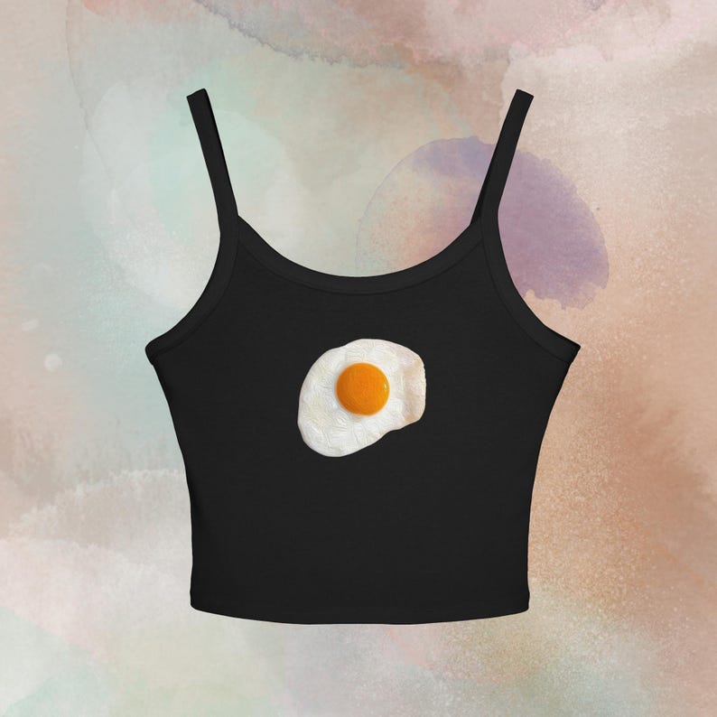May include: Black spaghetti strap crop top featuring a fried egg graphic. The egg has a bright yellow yolk and a white, textured egg white. The top is a casual, sleeveless style.