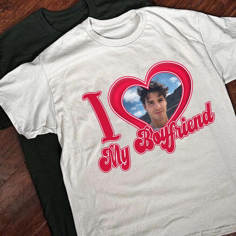 May include: White t-shirt with a red heart graphic containing a photo of a person. The words "I My Boyfriend" are printed in red script. A black t-shirt is partially visible in the background.