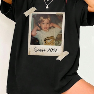 May include: Black t-shirt with a vintage photograph of a young child. The photo is attached with tape and has the text "Senior 2026". The child is wearing a t-shirt with the text "Pittsburgh Pirates".