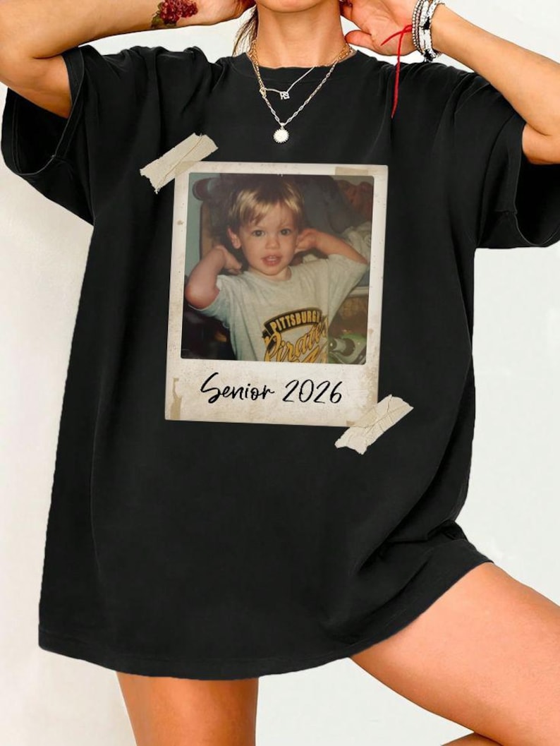 May include: Black t-shirt with a vintage photograph of a young child. The photo is attached with tape and has the text "Senior 2026". The child is wearing a t-shirt with the text "Pittsburgh Pirates".