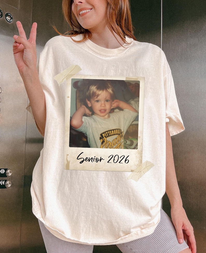 May include: Off-white t-shirt with a Polaroid-style photo of a child attached. The photo has the text "Senior 2026" beneath it. The child in the photo is wearing a shirt with the words "Pittsburgh Pirates".