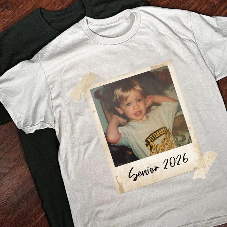 May include: White t-shirt with a vintage Polaroid photo of a young child, taped on with pieces of tape. The photo has the text "Senior 2026". A black t-shirt is partially visible in the background.