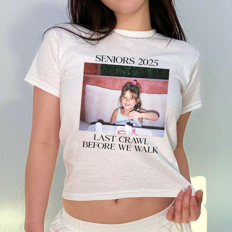 Op de afbeelding: Wit T-shirt met een foto van een jong meisje en de tekst "Seniors 2025" en "Last Crawl Before We Walk".