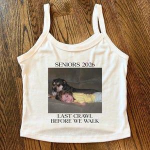 May include: White tank top with a photo print of a child and a dog. The photo is framed and includes the text "SENIORS 2026" and "LAST CRAWL BEFORE WE WALK". The tank top has a scoop neck and thin straps.