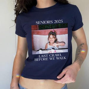 Op de afbeelding: Een kort, donkerblauw T-shirt met de tekst "SENIORS 2025" en "LAST CRAWL BEFORE WE WALK". In het midden van het shirt staat een foto van een jong meisje.