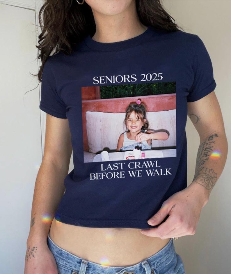 Op de afbeelding: Een kort, donkerblauw T-shirt met de tekst "SENIORS 2025" en "LAST CRAWL BEFORE WE WALK". In het midden van het shirt staat een foto van een jong meisje.