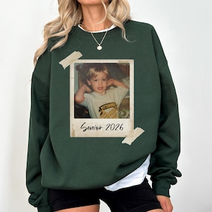 May include: Dark green sweatshirt featuring a vintage photo graphic. The photo is of a young child and is taped to the sweatshirt. The text "Senior 2026" is printed below the photo.