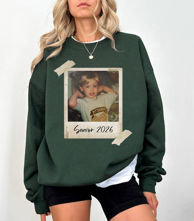 May include: Dark green sweatshirt featuring a vintage photo graphic. The photo is of a young child and is taped to the sweatshirt. The text "Senior 2026" is printed below the photo.