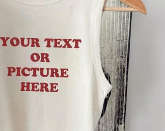 May include: Cream-colored tank top with the words "YOUR TEXT OR PICTURE HERE" printed in red. The top is sleeveless and hangs on a wooden hanger. The background is a textured, neutral color.