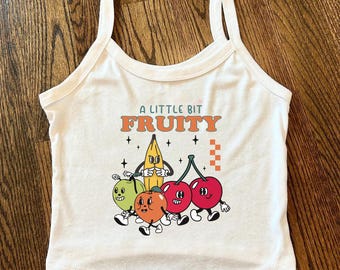 Può includere: Canotta bianca con la scritta "A Little Bit Fruity" e una grafica a cartoni animati di una banana, ciliegie, un'arancia e una mela verde. La canotta ha uno scollo tondo e spalline sottili.
