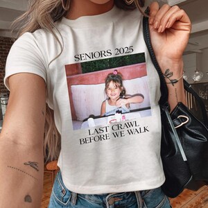 Op de afbeelding: Wit T-shirt met de tekst "Seniors 2025" en "Last Crawl Before We Walk". Een foto van een jong meisje is op het shirt gedrukt.
