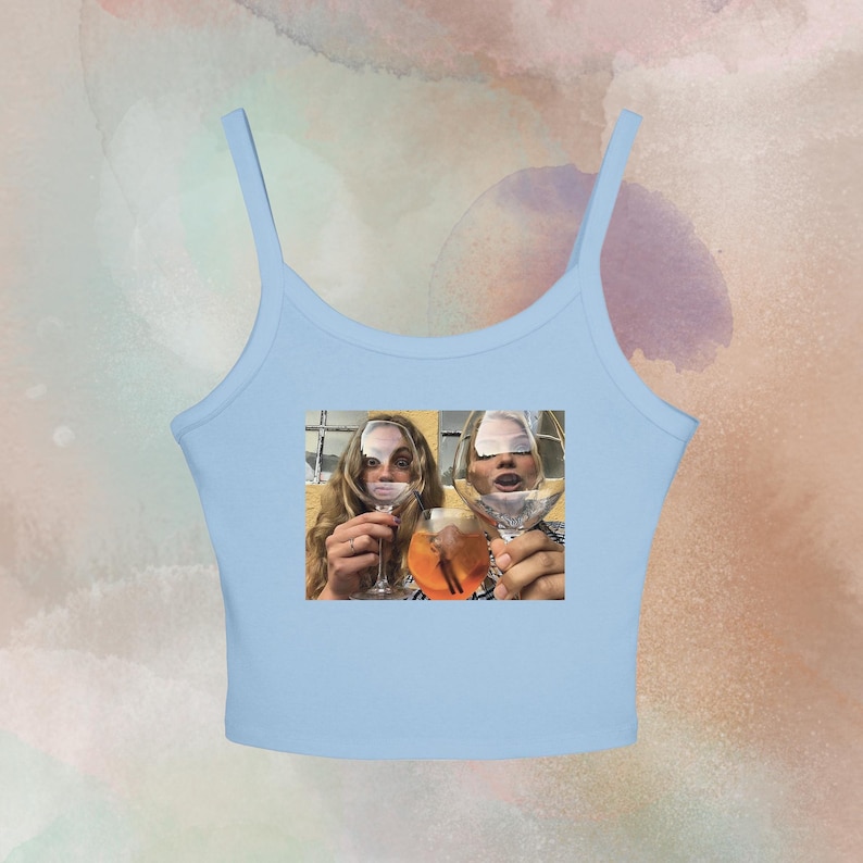 May include: Light blue tank top with a photo print of two people holding cocktail glasses. The image shows two women with their faces partially obscured by the glasses, filled with orange and clear liquids. The tank top has thin straps.