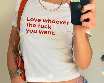 May include: White crop top with the text "Love whoever the fuck you want" in red. The shirt is paired with denim shorts and a small orange shoulder bag. A gold chain necklace is also visible.