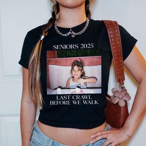Op de afbeelding: Zwart getailleerd T-shirt met een witte grafische print. De grafiek toont een foto van een jong meisje met de tekst "Seniors 2025" en "Last Crawl Before We Walk".