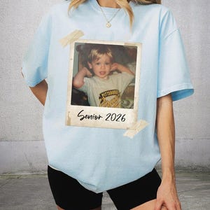 May include: Pale blue t-shirt featuring a retro photograph print. The photo shows a young child and is labelled "Senior 2026". The shirt is taped with pieces of beige tape.