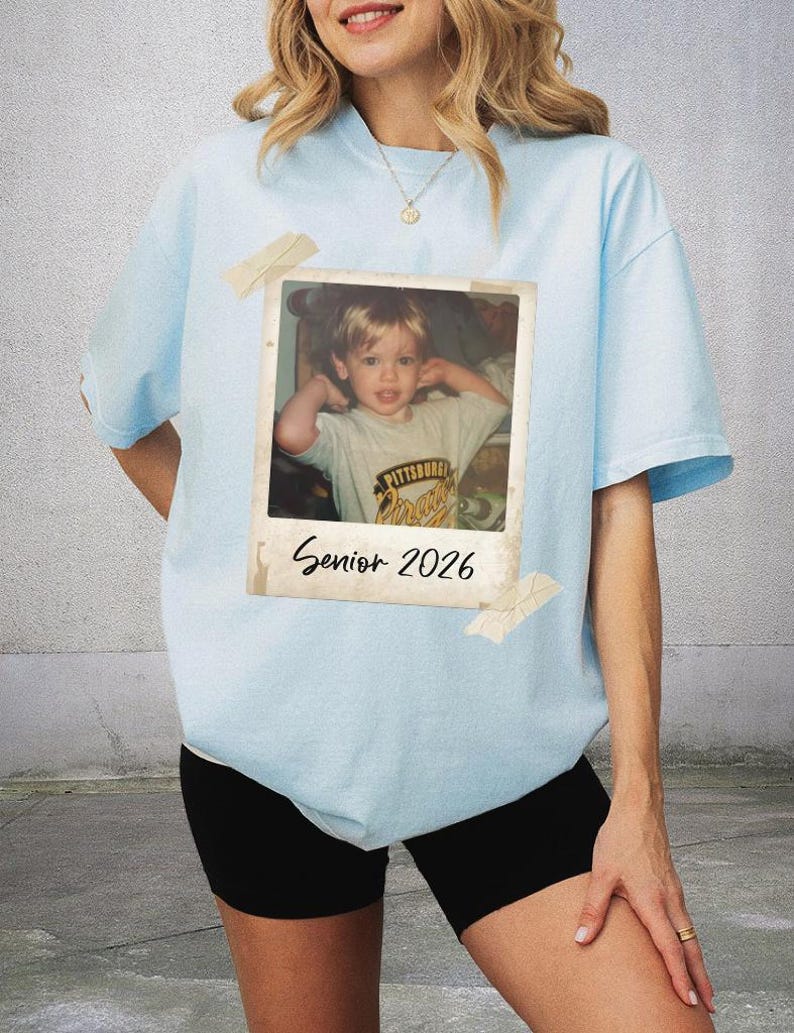 May include: Pale blue t-shirt featuring a retro photograph print. The photo shows a young child and is labelled "Senior 2026". The shirt is taped with pieces of beige tape.