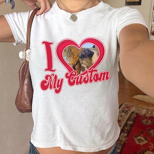 Peut inclure: T-shirt blanc avec l'inscription "I My Custom" en rouge, avec un cœur rouge contenant une photo d'un chien et d'une personne. Le t-shirt a des manches courtes.