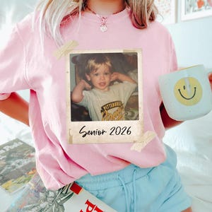 May include: A pale pink t-shirt with a retro photograph attached. The photo features a young child and the text "Senior 2026". A white mug with a yellow smiley face is held.