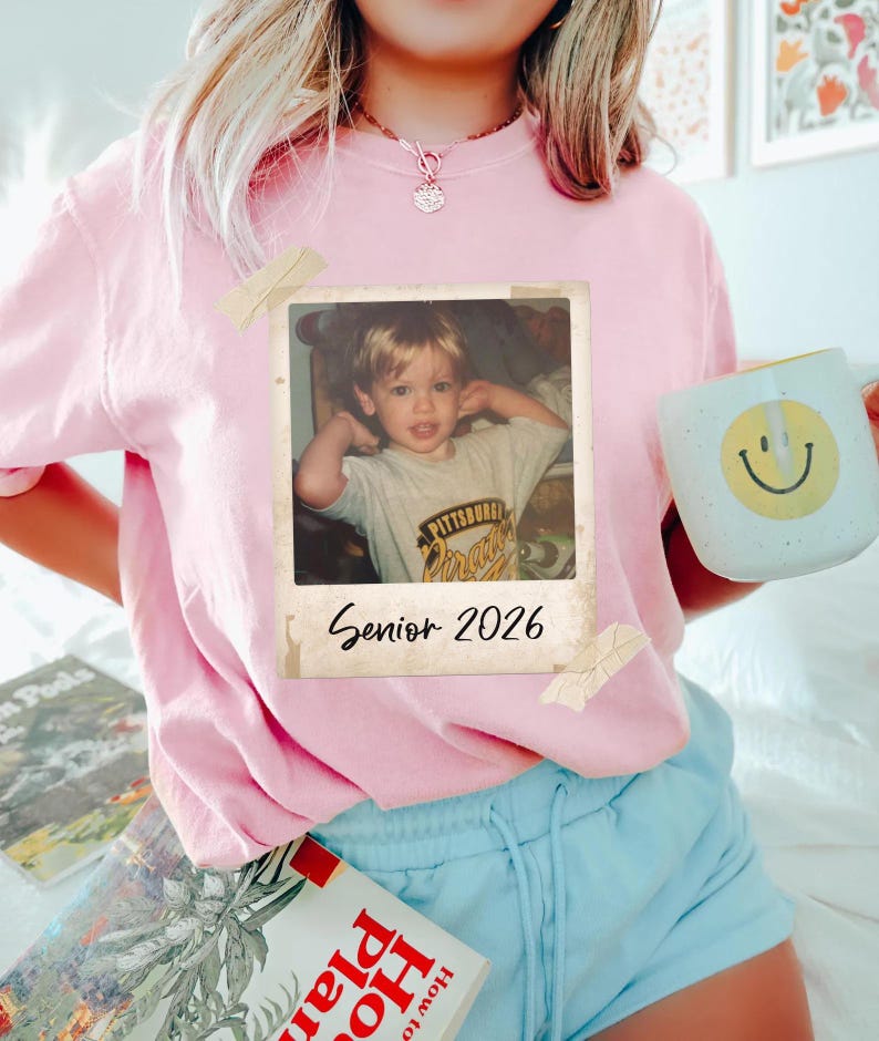 May include: A pale pink t-shirt with a retro photograph attached. The photo features a young child and the text "Senior 2026". A white mug with a yellow smiley face is held.