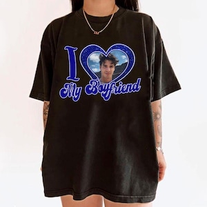 May include: Black t-shirt with a blue heart graphic containing a photo of a person. The text "I My Boyfriend" is written in blue script below the heart. The shirt has short sleeves and a crew neck.