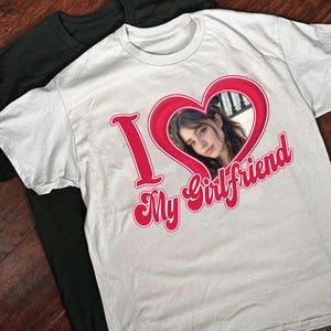 May include: A white t-shirt with a red heart design and the text "I love my girlfriend" in a red retro font. A woman's face is visible inside the heart.