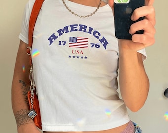 May include: White, short-sleeved, cropped t-shirt featuring the word "AMERICA" in blue arched letters, the American flag, and the text "17 76 USA" in blue.