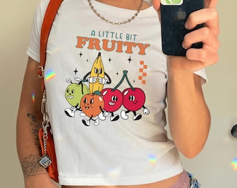 May include: White crop top with the text "A Little Bit Fruity" in orange and green. The shirt features a cartoon illustration of a banana, cherries, orange, and a green fruit with faces and arms.